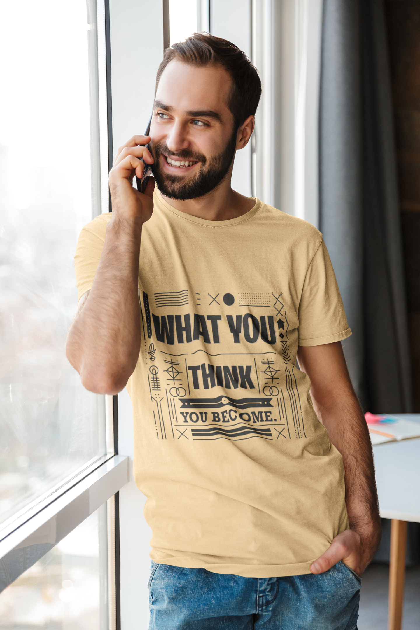 Man wearing a beige t-shirt with motivational text, talking on a phone indoors.