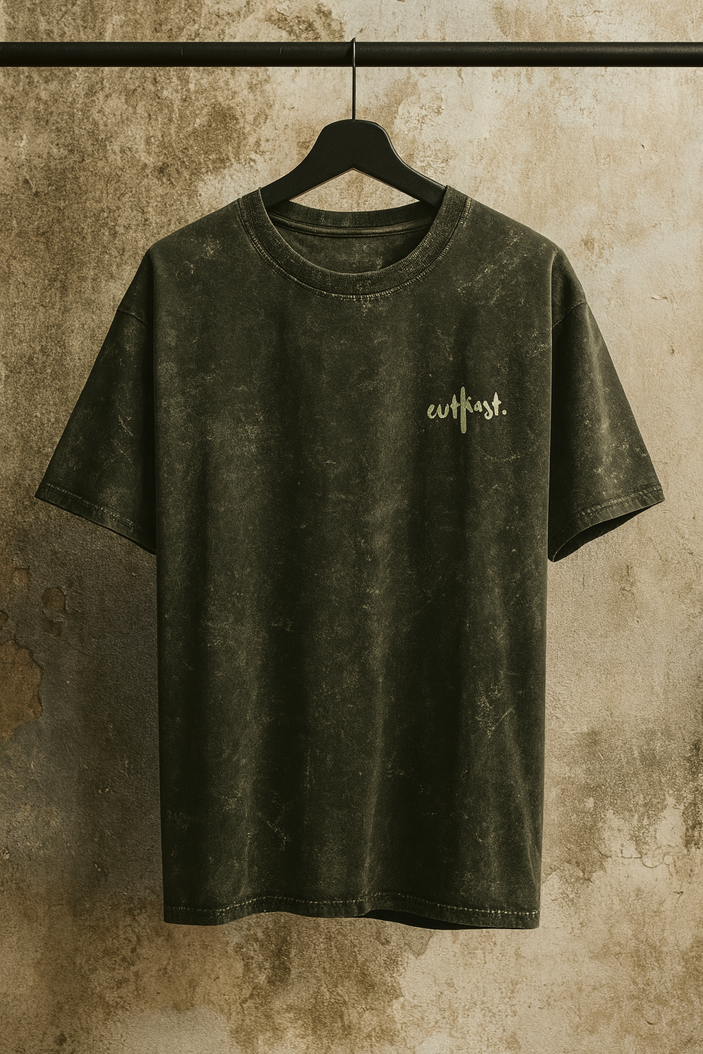 Dark Green acid-wash t-shirt with “outkast.” logo on chest, hanging on a black hanger against a rustic concrete wall.