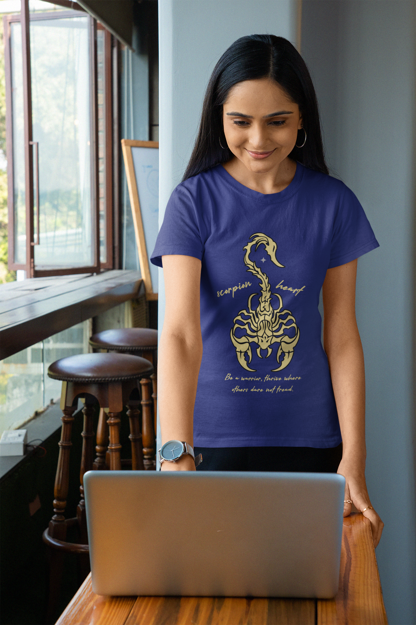 Woman wearing a blue t-shirt with a gold scorpion design, sitting at a table with a laptop.