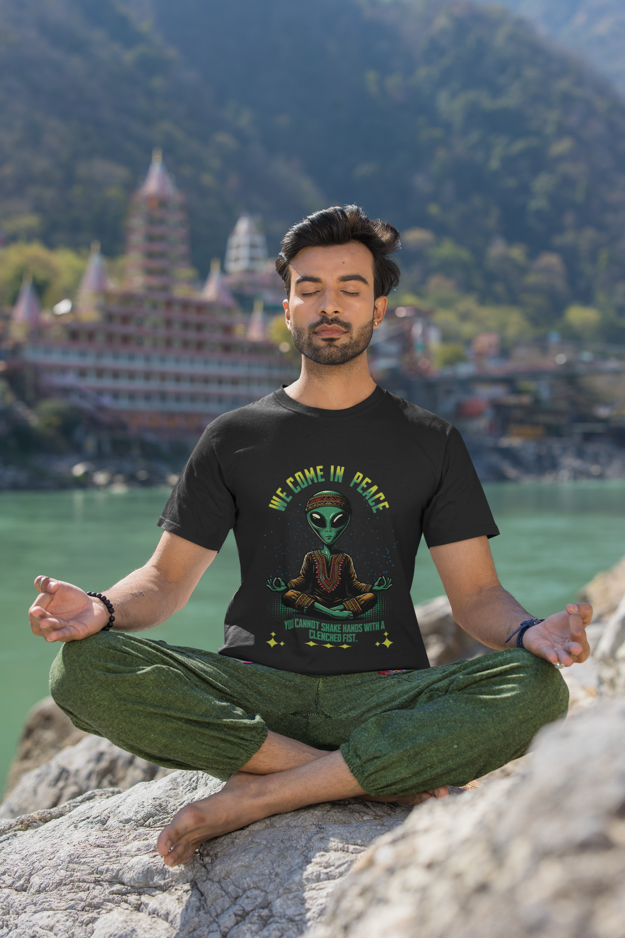 Man meditating on a rock with a scenic background and a t-shirt with an alien design.
