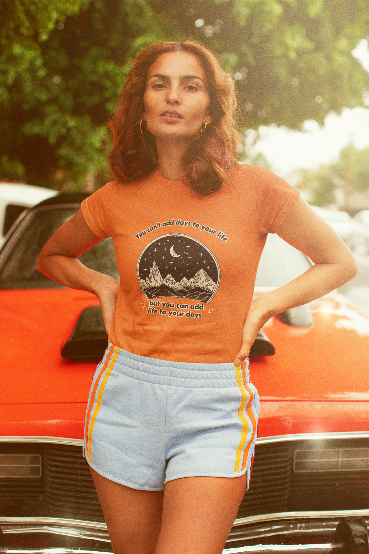Woman wearing an orange t-shirt with a graphic design, standing in front of a vintage car.
